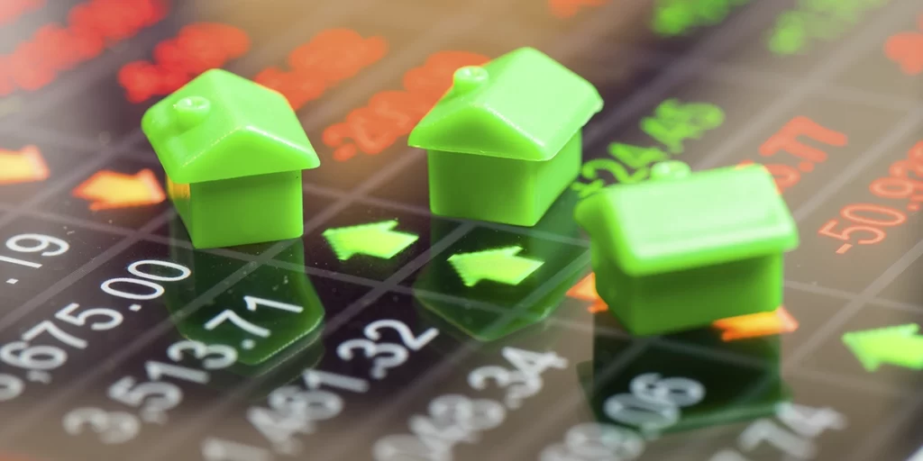 Índice Imobiliário Índice Imobiliário: A imagem mostra três peças verdes de um brinquedo, que parecem ser peças de uma casa, sobre um fundo com gráficos financeiros. O fundo apresenta gráficos de desempenho de ações ou índices financeiros, com ícones de setas vermelhas e verdes, indicando movimentos de preço. As casas em miniatura são representação visual simbólica do mercado financeiro, possivelmente sugerindo investimentos em imóveis ou ações de empresas relacionadas ao setor imobiliário.