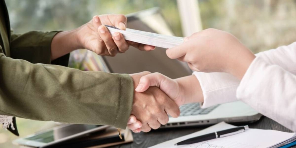 Pagamento de comissões Pagamento de comissões: close num aperto de mãos entre duas pessoas. Com as outras mãos, uma entrega um papel a outra.