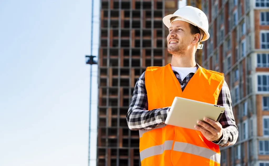 Anotação de Responsabilidade Técnica Anotação de Responsabilidade Técnica: A imagem mostra um homem vestindo um capacete branco e um traje de segurança amarrado, que inclui uma jaqueta de segurança laranja e um colete de proteção. Ele está segurando um tablet e parece estar olhando para algo acima dele, possivelmente um edifício em construção. O cenário ao fundo é um dia claro com um edifício em construção ao fundo, indicando que o homem pode ser um trabalhador de construção ou um engenheiro de projeto.