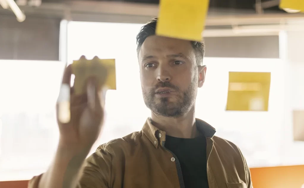 Anotação de Responsabilidade Técnica Anotação de Responsabilidade Técnica: A imagem mostra um homem de cabelo curto e barba pequena, vestindo uma camisa de manga curta e um casaco de manga longa. Ele está segurando uma folha amarrada ao dedo e olhando para ela com uma expressão pensativa. Há outras folhas amarradas ao dedo suspensas ao redor dele, sugerindo uma atividade de organização ou planejamento. O fundo é desfocado, mas parece ser um ambiente interno com uma janela ou porta aberta, permitindo uma luz natural que ilumina a cena.