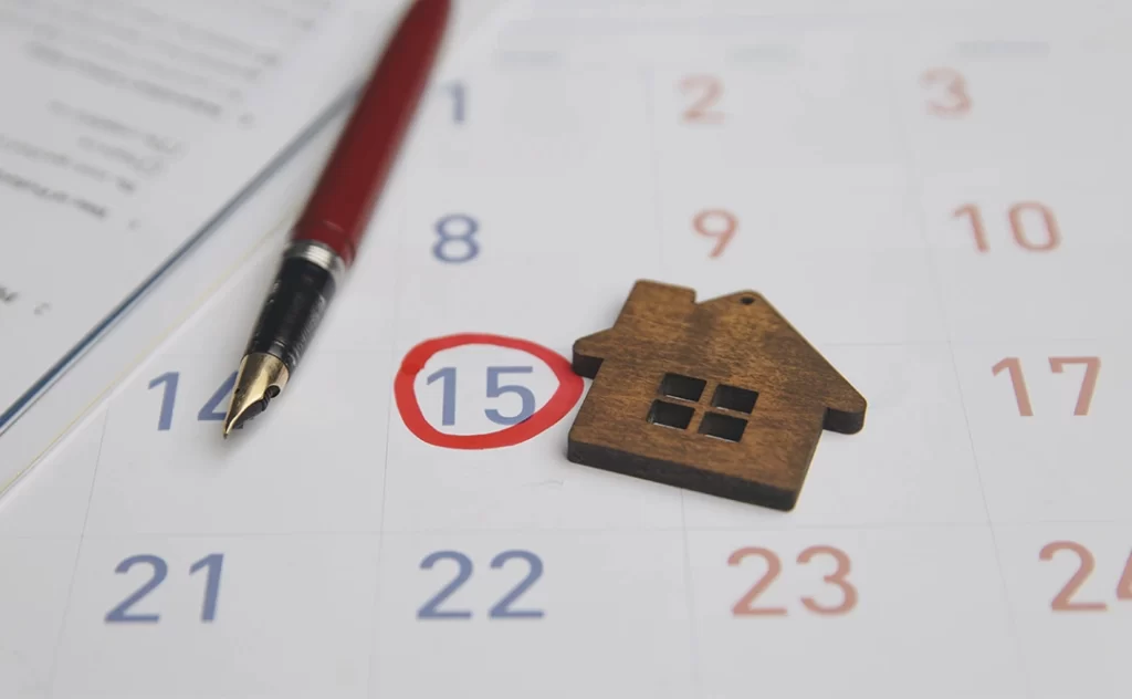 Calendário imobiliário Calendário imobiliário: A imagem mostra um calendário de mês com o dia 15 marcado com um círculo vermelho. Ao lado do calendário, há um ponto de escrita preto e um pequeno logotipo de uma casa, que parece ser um marcador de calendário ou um adesivo decorativo. O calendário está sobre um papel com texto, e a data marcada pode indicar um evento importante, como uma data de mudança ou uma data de contrato de aluguel.