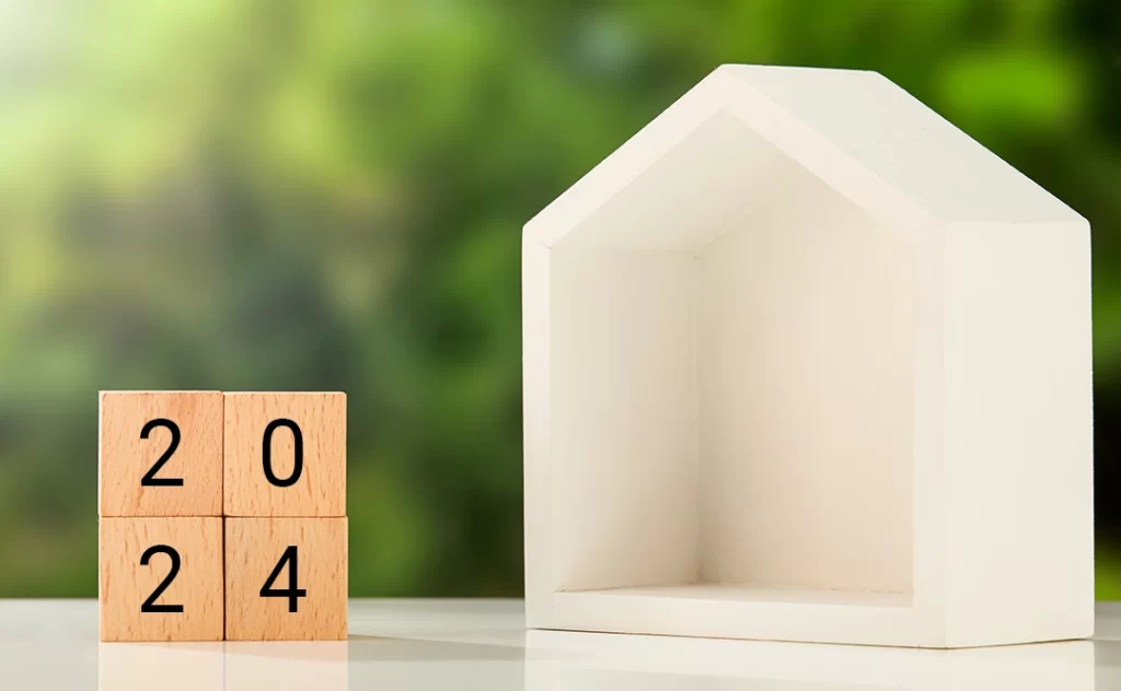 Calendário imobiliário Calendário imobiliário: A imagem mostra um cubo de madeira com o número "24" escrito em preto, ao lado de um modelo de casa de madeira de cor branca. Ambos estão sobre uma superfície branca, e ao fundo, há uma área de vegetação com tons de verde suavemente desfocada.