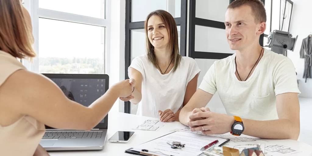 Explique como funciona o pré-atendimento Explique como funciona o pré-atendimento: A imagem mostra uma equipe de pessoas sentadas em uma mesa em um ambiente de escritório. Uma pessoa está dando a mão para outra pessoa, enquanto uma terceira pessoa olha. As pessoas estão todos vestidas com roupas de negócios e parecem estar discutindo algo. Existe um laptops na mesa em frente delas e um pedaço de papel na mesa em frente da pessoa que está dando a mão. A sala está bem iluminada e parece ser moderna e profissional.