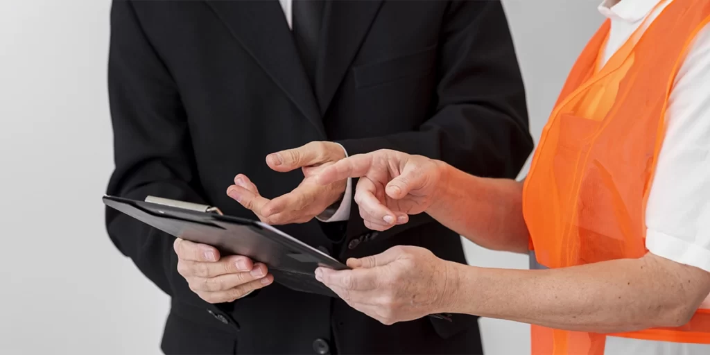 Gestão de mão-de-obra Gestão de mão-de-obra: A imagem mostra duas pessoas em um ambiente interno. A pessoa à esquerda está vestindo um terno preto e parece estar olhando para uma tabela ou um dispositivo que a outra pessoa está apresentando. A segunda pessoa, à direita, está vestindo uma jaqueta amarla e parece estar explicando ou mostrando algo ao primeiro indivíduo. Ambas as pessoas estão de pé e parecem estar em um contexto de trabalho ou apresentação.