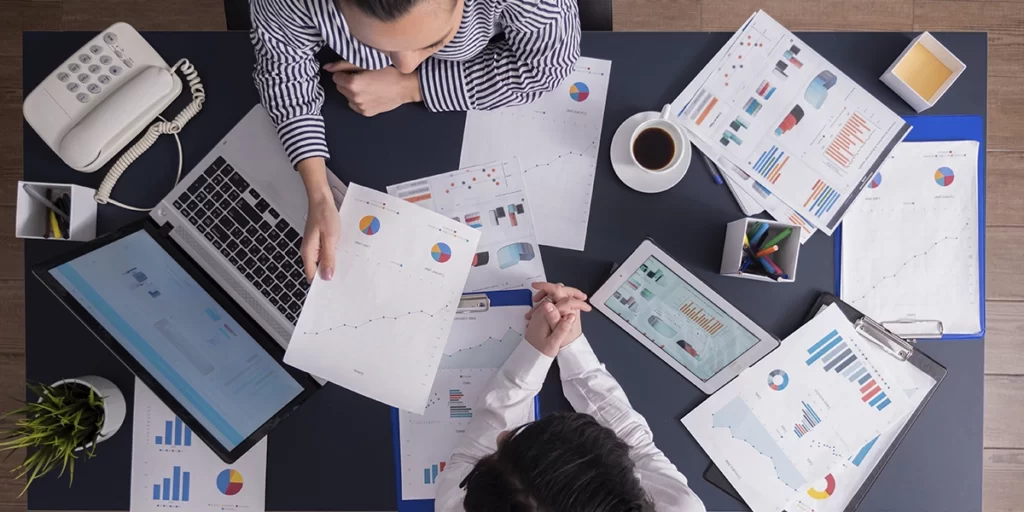 Plano de Negócio Plano de Negócio: A imagem mostra duas pessoas sentadas em frente a um grande tabela preto, trabalhando em um ambiente de negocios. Eles estão examinando documentos e gráficos, que parecem ser relatórios de negócios ou estatísticas. Há vários itens de escritório em cima do tabela, incluindo um telefone, um laptop, um tablet, um par de óculos, um par de canetas e um copo de café. A atmosfera é organizada e profissional.