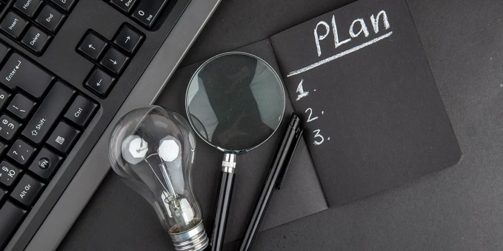 Plano de Negócio Plano de Negócio: A imagem mostra uma escrivaninha preta com o texto "Plan" escrito em branco no topo. Abaixo do texto, há três números: "1.", "2." e "3.", também escritos em branco. Ao lado da escrivaninha, há um teclado preto com alguns botões visíveis. Ao lado do teclado, há um par de óculos de armação preta e lentes brancas, e um par de lâmpadas de armação preta e vidro claro. A lâmpada está parcialmente dentro da escrivaninha, e o par de óculos está parcialmente dentro da lâmpada. A imagem tem um estilo moderno e parece ser uma representação visual de planejamento ou ideias.