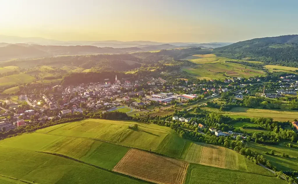 Terrenista Terrenista: A imagem mostra uma paisagem rural com uma vista aérea. Há uma cidade com edifícios de arquitetura tradicional, rodeada por campos de grama verde e áreas de cultivo. Ao fundo, há uma série de colinas e montanhas, sugerindo uma área de terra baixa ou uma região costeira. O sol está baixo no horizonte, indicando que pode ser perto do horário do pôr do sol ou do nascer do sol, o que dá à cena um aspecto tranquilo e pitoresco.