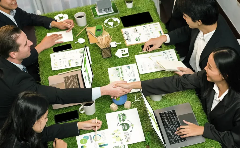 Terrenista Terrenista: A imagem mostra uma cena de negociações ou reunião de negócios em um ambiente moderno e casual. Há cinco pessoas sentadas em volta de uma mesa com grama artificial verde, sugerindo um ambiente de trabalho eficiente e ambiente. Dois homens estão se dando um abraço de mão, indicando um acordo ou um momento de celebração. As outras pessoas estão engrossadas em documentos e dispositivos, como laptops e celulares, sugerindo que estão trabalhando ou discutindo negócios. A mesa está bem organizada com notas, apresentações e outros materiais relacionados ao trabalho. A atmosfera é profissional, mas com um toque de casualidade, possivelmente indicando uma reunião de negócios mais informal ou uma equipe que trabalha de forma colaborativa e descontraída.