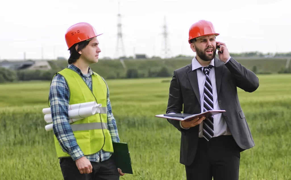 Terrenista Terrenista: A imagem mostra dois homens em um ambiente ao ar livre, possivelmente um campo ou uma área de construção. O homem à esquerda está vestindo uma jaqueta amarilla de segurança, um capacete vermelho e uma camisa de manga curta com padrões. Ele parece estar olhando para o lado, possivelmente atento a algo fora da cena. O homem à direita está usando um terno preto, uma camisa branca com uma gravata preta e branca, e também um capacete vermelho. Ele está falando em um telefone celular e parece estar segurando algum material, talvez um livro ou um folheto. Ambos parecem estar trabalhando juntos em um projeto ou uma tarefa.