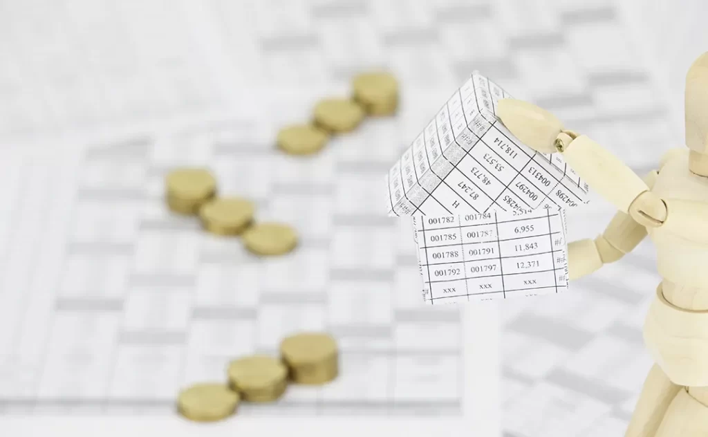 Venda de Loteamento A imagem mostra uma figura de boneco de madeira, que parece ser um modelo de um humano, com um braço estendido para a direita. No braço, há uma pequena estrutura que parece ser um modelo de uma casa ou edifício, com várias etiquetas ou placas de identificação. Ao longo do braço, há uma série de moedas ou peças de metal, que parecem ser colocadas em uma ordem específica. Ao fundo, há uma superfície com uma grade ou quadriculado, que pode ser uma representação de um plano de projeto ou um mapa.