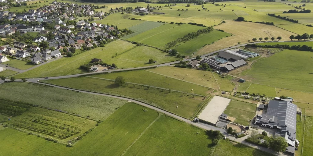 compra de lotes: Esta imagem mostra um paisagem rural com uma pequena aldeia no primeiro plano. A aldeia é cercada por campos verdes e árvores, e várias casas e edifícios são visíveis no horizonte. O céu é claro e azul, e há algumas nuvens no horizonte. A imagem foi tirada de uma altura elevada, oferecendo uma visão aérea da paisagem.