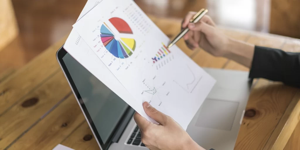 diagrama de ishikawa diagrama de ishikawa: A imagem mostra uma mão de um indivíduo sentado em frente a um laptop aberto. A mão está segurando uma folha de papel com gráficos e dados, possivelmente um relatório ou um estudo estatístico. A outra mão está segurando um caneta, sugerindo que o indivíduo pode estar anotando ou estudando os dados na folha. O ambiente parece ser um espaço de trabalho ou escritório, com uma mesa de madeira e uma cadeira ao fundo.