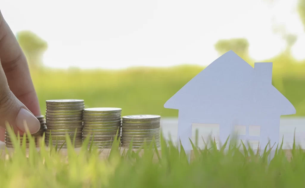 investir em lotes investir em lotes: imagem de uma mão empilhando moedas numa grama. As moedas estão em destaque e, ao lado, uma casa de papel.