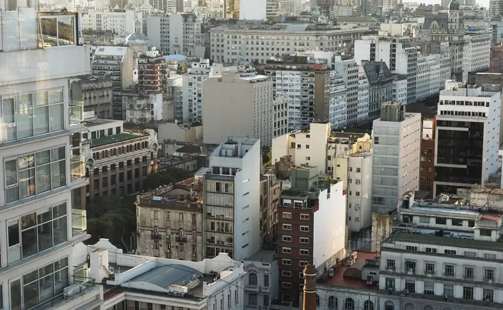 Urbanização e construção civil Urbanização e construção civil: A imagem mostra uma vista aérea de uma área urbana densa. Há uma variedade de edifícios de arquitetura moderna e histórica, com vários andares e cores que variam de brancos e claros a tons mais escuros. A paisagem é dominada por edifícios residenciais e comerciais, com alguns espaços abertos que parecem ser parques ou áreas de terra nua. A cena transmite uma sensação de grandeza e complexidade, com a cidade se estendendo até o horizonte.