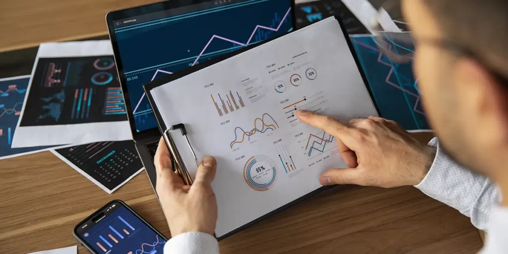 CRM na gestão de vendas de lotes CRM na gestão de vendas de lotes: A imagem mostra uma pessoa sentada em frente a um computador, com uma folha de papel aberta em frente a ela. A folha de papel contém gráficos e diagramas que parecem ser estatísticas ou dados de negócios. A pessoa está usando óculos e parece estar examinando ou explicando os gráficos. No fundo, há outros documentos e um celular com uma tela aberta que também mostra gráficos semelhantes aos do papel. A cena sugere um ambiente de trabalho ou estudo, possivelmente relacionado a análises de dados ou planejamento estratégico.