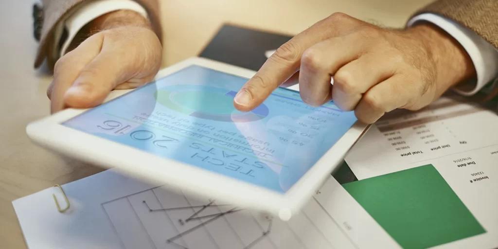 CRM na gestão de vendas de lotes CRM na gestão de vendas de lotes: A imagem mostra uma mão humana, provavelmente de um adulto, usando um tablet. A mão está pressionando o touchscreen do tablet, que está aberto e exibe uma interface de usuário com gráficos e texto. O tablet está sobre um suporte que parece ser um bloco de notas ou um livro, e há outros documentos ou folhas de papel ao redor, sugerindo um ambiente de trabalho ou estudo. A mão está vestida com um cinto de camisa branca, o que pode indicar um contexto profissional ou formal. A imagem é uma foto real e parece ser tirada de cima, focando na interação do usuário com o tablet.