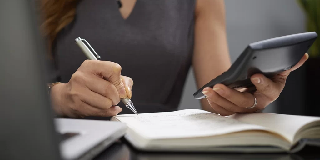 como funciona o imposto de renda para empresas: A imagem mostra uma pessoa sentada ao redor de uma mesa, focada em trabalhar ou estudar. Ela está usando um smartphone e um caderno. O smartphone está aberto e parece estar sendo usado para algum tipo de aplicativo ou software, possivelmente relacionado a estudo ou trabalho. A pessoa está escrevendo ou tocando o caderno com um lápis, sugerindo que ela está tomando notas ou trabalhando em algum projeto. Ela está vestindo uma blusa preta e parece estar em um ambiente de escritório ou estudo, com uma planta ao fundo e uma janela parcialmente visível. A pessoa está usando um colar e parece estar concentrada em suas tarefas.