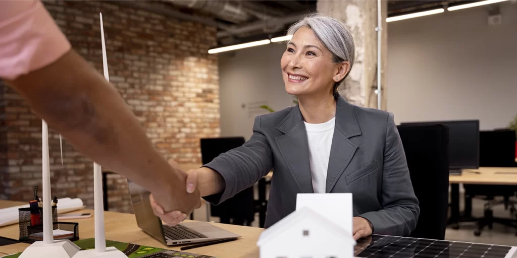 O que é um lead qualificado O que é um lead qualificado: A imagem mostra duas pessoas em um ambiente que parece ser um escritório moderno. A pessoa de cabelos grisalhos está sentada atrás de uma mesa, vestindo um terno azul e uma camisa branca. Ela está sorrindo e se estende para cumprimentar a outra pessoa, que está de pé e estende a mão para a sua. A pessoa de pé está vestindo uma camisa rosa e parece estar segurando algo na mão, mas não é possível ver o que é.
A mesa da pessoa sentada tem um laptop aberto, um modelo de casa branca e alguns outros objetos que parecem ser material de trabalho. Há uma parede de ladrilhos ao fundo e uma janela com uma janela de vidro. O ambiente é iluminado naturalmente e parece ser um espaço de trabalho agradável e moderno.
