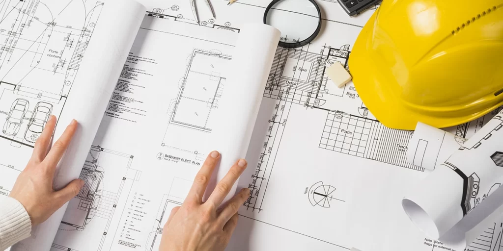 as built as built: A imagem mostra duas mãos de uma pessoa abrindo e examinando um livro de projetos arquitetônicos. Ao fundo, há outros documentos de projeto e equipamentos relacionados ao setor de construção, como um capacete amarrado e um estetoscópio. A cena sugere um ambiente de trabalho de projeto de construção ou arquitetura.