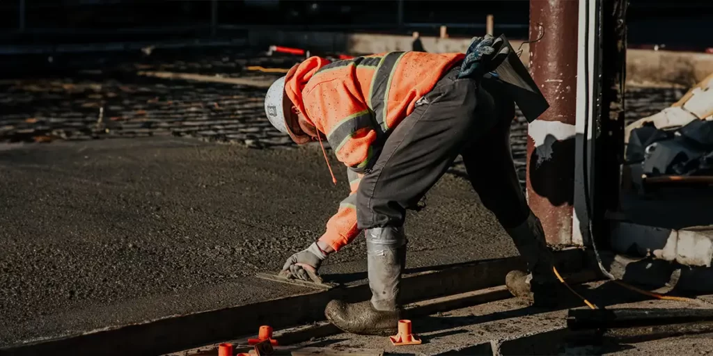 concreto usinado concreto usinado: A imagem mostra uma pessoa em um ambiente de construção. A pessoa está de costas para a câmera, com uma postura baixa, sugerindo que está trabalhando. Ela está vestindo um traje de segurança que inclui uma jaqueta amarronada e um capacete, indicando que está em um ambiente de trabalho onde a proteção é necessária. A jaqueta tem detalhes de segurança, como linhas amarelas e vermelhas, que são comuns em trajes de segurança para aumentar a visibilidade. A pessoa está usando calças pretas e parece estar usando algum tipo de protetor para as mãos, possivelmente para segurança contra ferramentas ou materiais.