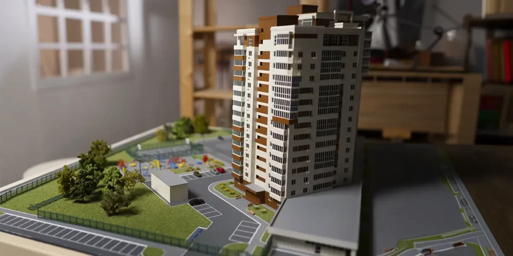 empreendimentos mistos empreendimentos mistos: A imagem mostra uma representação em miniatura de um complexo residencial. Há um edifício de apartamentos de vários andares, com uma fachada que combina cores brancas e pretas.
No piso inferior, há um parque infantil com equipamentos de brinquedo,
O complexo tem um estacionamento aberto com vários espaços para carros. Há também uma área de passeio com grama e plantas decorativas. Ao lado do edifício, há uma área de entrada com uma porta e uma área de espera.
A representação é feita em escala, possivelmente para ilustrar o design do complexo antes de sua construção ou para uso em um modelo de arquitetura. A imagem é uma foto da cena modelada, e não é uma foto realista do complexo em si.