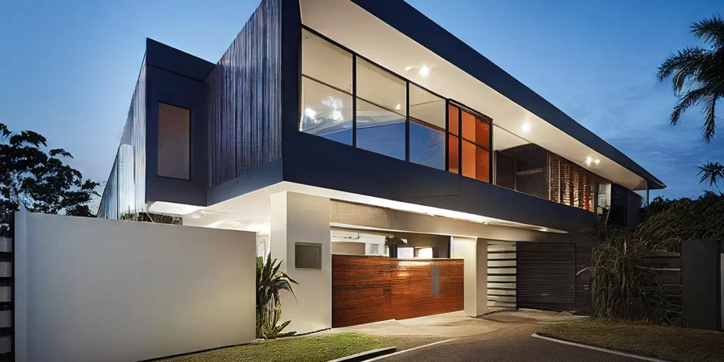 Imagem de uma casa grande e moderna com um andar elevado e muitas janelas de vidro. No andar debaixo vemos uma garagem subterrânea com o portão de madeira fechado.