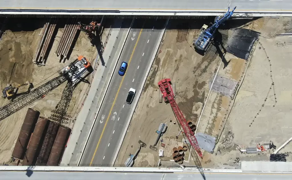 logística de canteiro de obras logística de canteiro de obras: Esta é uma visão aérea de um local de construção com um crante grande e várias peças de equipamentos no chão. O crante está levando uma grande peça de aço para o lugar, enquanto trabalhadores usando capacetes e roupas de trabalho estão ao redor dele. Existem também várias outras peças de equipamentos no chão, incluindo um grande excavador e um caminhão de lixo. O céu é claro e azul, e há algumas árvores no fundo.