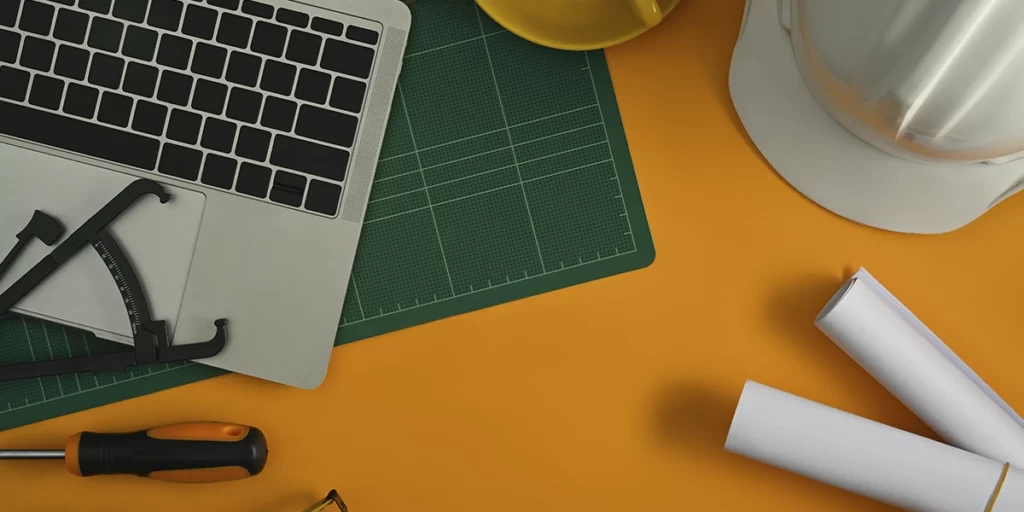 materiais inovadores materiais inovadores: A imagem mostra uma superfície amarela com vários itens relacionados a trabalhos de construção ou manutenção. Há um laptop aberto com o teclado visível, um capacete, papeis de plantas de imóvel, uma chave de fenda.