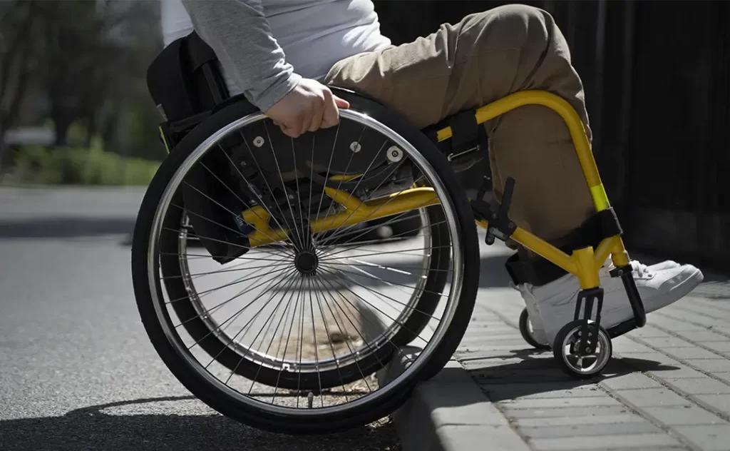 nbr 9050 atualizada nbr 9050 atualizada: A imagem mostra o corpo de uma pessoa em uma cadeira de rodas com. A pessoa está vestindo uma camisa branca e calça bege. A cadeira de rodas é amarela com listras pretas. O fundo é uma rua da cidade com árvores e edifícios visíveis no horizonte.