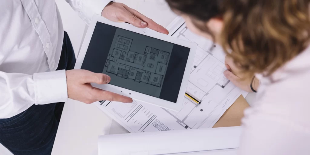 sistema de gestão para construtoras sistema de gestão para construtoras: A imagem mostra duas pessoas em um ambiente que parece ser um escritório ou uma sala de trabalho. A pessoa à frente está segurando um tablet de tela grande, que está exibindo um plano de uma casa. A outra pessoa está sentada ao fundo, com uma folha de papel ou plano de projeto em frente a ela, e parece estar olhando para o tablet. Ambas parecem estar trabalhando em um projeto de arquitetura ou design de interiores, possivelmente discutindo detalhes do projeto ou comparando o plano digital com o plano impresso. Ambas estão vestidas com camisetas brancas, sugerindo um ambiente casual de trabalho.