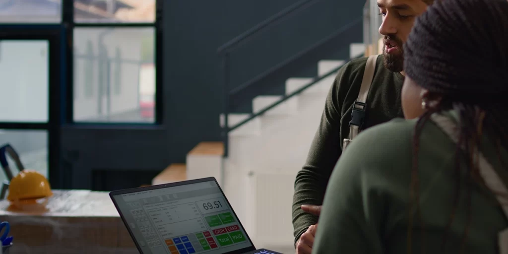 sistema de gestão para construtoras sistema de gestão para construtoras: A imagem mostra duas pessoas em um ambiente interno, possivelmente uma oficina ou um escritório. A pessoa à frente está usando uma camisa vermelha e parece estar olhando para um laptop aberto em frente a ele. O laptop está exibindo uma tela de um sistema de pagamento, com informações como "69,51" e botões como "SALVAR", "CANCELAR", "PAGAR", e "SALDO". A outra pessoa, que está de costas para a câmera, está vestindo uma camisa verde e parece estar participando da conversa ou da tarefa em andamento. Ambos parecem estar engrossados em algum tipo de trabalho ou discussão relacionada ao conteúdo do laptop. O ambiente é iluminado com luz artificial e há uma janela ao fundo que dá uma visão parcial do exterior.