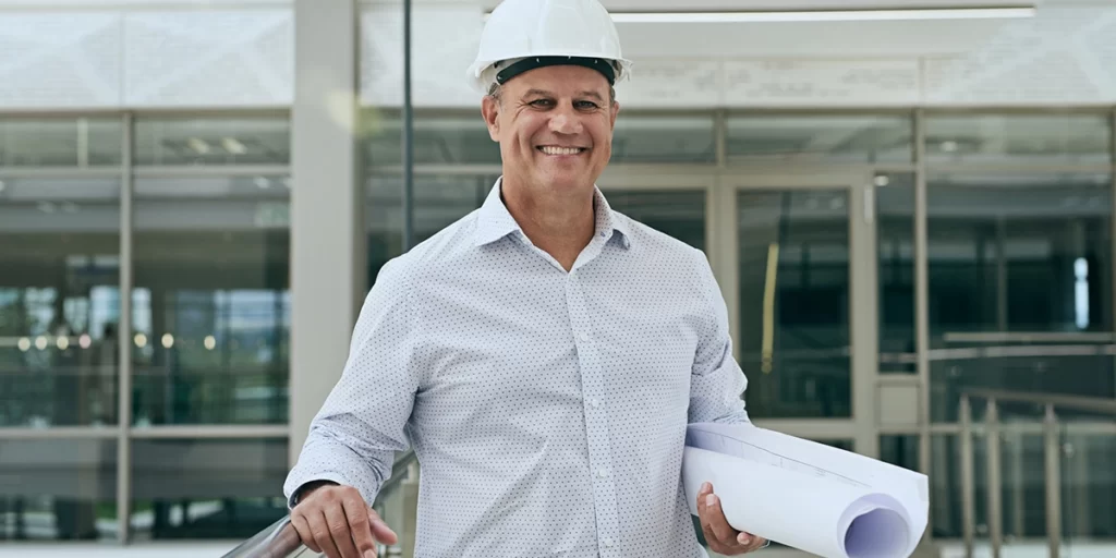 software de gestão para construtoras software de gestão para construtoras: A imagem mostra um homem usando um capacete branco e segurando um conjunto de planilhas. Ele está em uma escada levando para um prédio com grandes janelas e um design moderno. O homem está sorrindo e parece feliz. O fundo é um prédio moderno com janelas de vidro e uma fachada branca. A imagem está bem iluminada e as cores são vibrantes e vivas. A atmosfera geral da imagem é alegre e otimista.