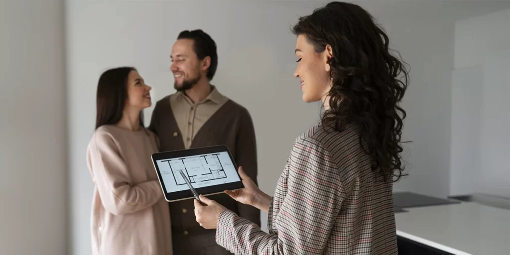 CRM para imobiliária CRM para imobiliária: A imagem mostra três pessoas em um ambiente interno que parece ser uma sala de design ou uma oficina de arquitetura. A pessoa de frente, uma mulher com cabelos longos e escuros, está segurando um tablet e parece estar mostrando algo aos outros dois indivíduos. Eles estão olhando para o tablet, sugerindo que estão examinando um projeto ou plano. A mulher está vestindo uma blusa com padrões e parece estar engrossada em o que está sendo mostrado no tablet. O homem ao lado dela está vestindo uma camisa e um cardigan e parece estar interessado no conteúdo do tablet. A terceira pessoa, uma mulher mais jovem, está segurando o braço do homem e também está olhando para o tablet. Todos parecem estar em um ambiente de trabalho ou de projeto, e a atmosfera é casual e colaborativa.