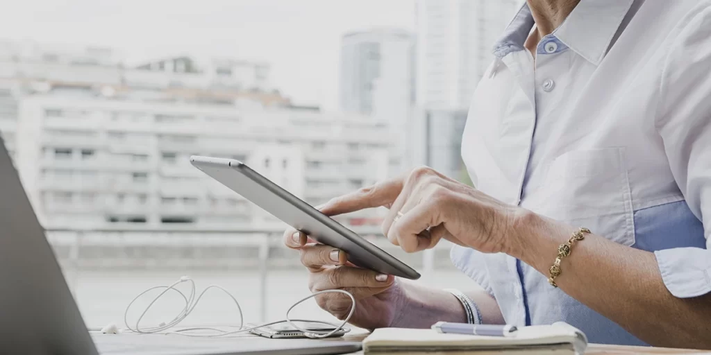 CRM para imobiliária CRM para imobiliária: A imagem mostra uma pessoa sentada em frente a um laptop. A pessoa está usando um tablet com um dedo, possivelmente para tocar ou desenhar no ecrã. Ela está vestindo uma camisa azul e parece estar trabalhando ou desenvolvendo algo no tablet. Há um par de fones de ouvido de cabo curto ao lado do laptop, sugerindo que ela pode estar usando áudio ou estar pronto para usar. Ao fundo, há uma vista de edifícios modernos, o que sugere que a cena pode estar acontecendo em um ambiente urbano, talvez em um escritório ou espaço de trabalho. A iluminação é suave e natural, indicando que pode ser um dia claro.