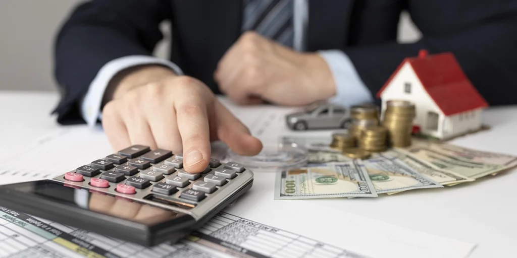 captação de recursos captação de recursos: A imagem mostra uma mão humana, provavelmente de um adulto, usando um cálculador de papel. A mão está pressionando o botão "+" do cálculador. Ao redor da mão e do cálculador, há dinheiro em espécie, moedas, e algumas peças de brinquedo que representam um carro e uma casa, sugerindo uma situação financeira ou de negociações. A mão está vestindo um colar e parece estar sobre um papel que pode ser um documento financeiro ou de contabilidade.