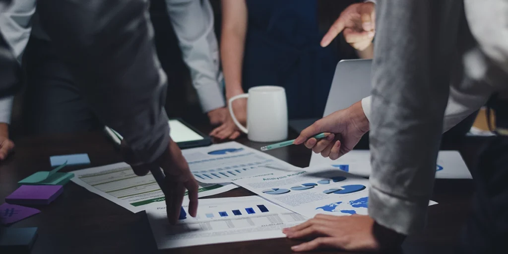 captação de recursos captação de recursos: A imagem mostra uma cena de trabalho em um ambiente de escritório. Há várias pessoas sentadas em volta de uma mesa, engrossadas em uma discussão ou análise de dados. A mesa está coberta com documentos, parecendo ser um relatório ou planilha com gráficos e dados em formato tabular.