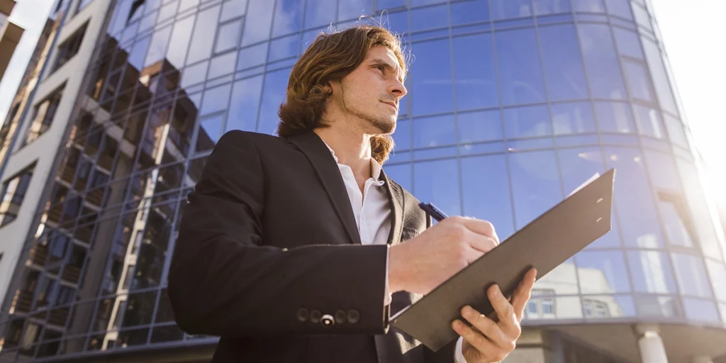 certificação edge certificação edge: imagem em contra plano de um homem usando terno e segurando uma planilha. A expressão dele é séria. Ao fundo um grande edifício espelhado.
