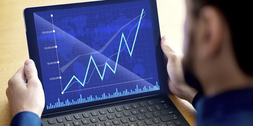 ciclos imobiliários ciclos imobiliários: A imagem mostra uma pessoa sentada em frente a um tablet com uma tela azul. A tela exibe gráficos e dados que parecem representar informações financeiras ou estatísticas, como gráficos de linha e barras. Há um aumento nos dados apresentados, indicado pela linha que se eleva ao longo do tempo. A pessoa está usando o tablet com ambas as mãos, o que sugere que ela está ativa e engajada com o conteúdo do dispositivo. O ambiente ao redor é um ambiente de escritório, com uma mesa e um teclado físico ao lado do tablet.