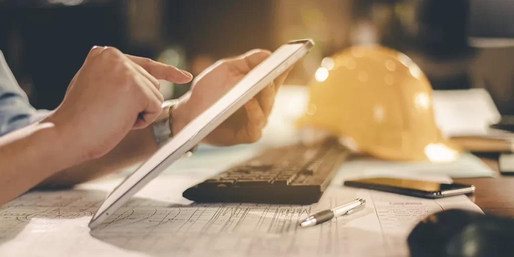 cronograma fisico e financeiro de obras cronograma fisico e financeiro de obras: A imagem mostra uma mão humana ao lado de um tablet de tela grande, que está aberto e colocado sobre um teclado. A mão está segurando o tablet, e parece estar a tocar ou interagir com o ecrã. Há um ambiente de escritório ao fundo, com uma luz suave que ilumina a parte superior do tablet e a mão, criando um efeito de profundidade de campo. Há também um capacete de construção ao fundo, sugerindo que a pessoa pode estar trabalhando em um projeto de construção ou estar em um ambiente de trabalho relacionado ao setor de construção. A imagem tem um filtro que dá uma sensação de calma e concentração.