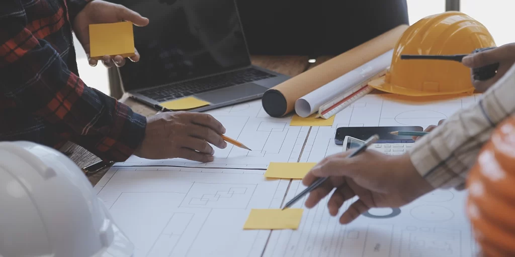 lean construction lean construction: A imagem mostra uma cena de trabalho em um ambiente que parece ser um escritório de engenharia ou construção. Há quatro pessoas sentadas em volta de uma mesa, todas vestidas com roupas amarelas, que são comumente usados para segurança em construção ou como indicação de trabalhadores de engenharia.