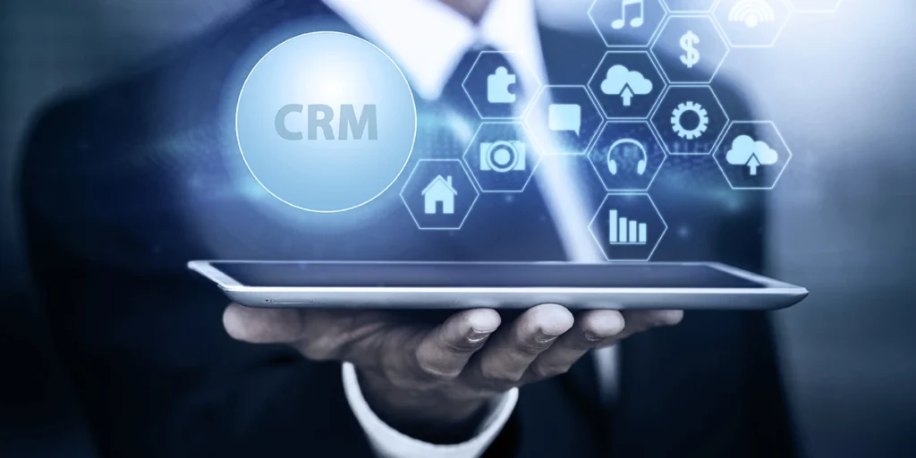 CRM para incorporadoras CRM para incorporadoras: close na mão de uma pessoa segurando um tablet. Da tela do tablet saem alguns ícones que representam conexão e relacionamento. Em destaque a sigla CRM.