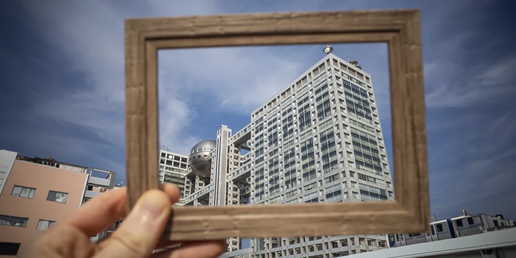 A imagem apresenta uma mão segurando uma moldura que está apontada para um prédio, fazendo com que ele fique em destaque.