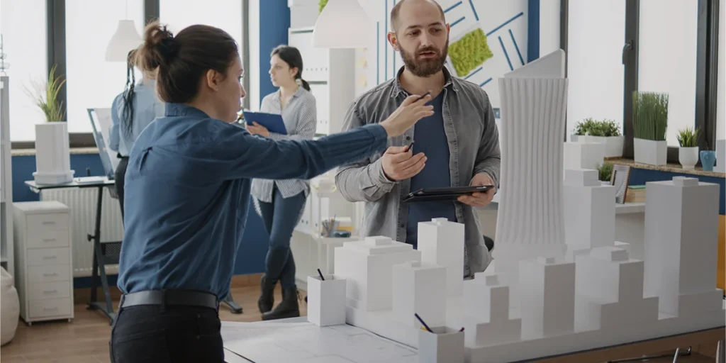 segmentação de clientes de construtoras segmentação de clientes de construtoras: imagem de duas pessoas conversando e apontando para uma maquete de edifício. Os dois estão num amplo escritório.