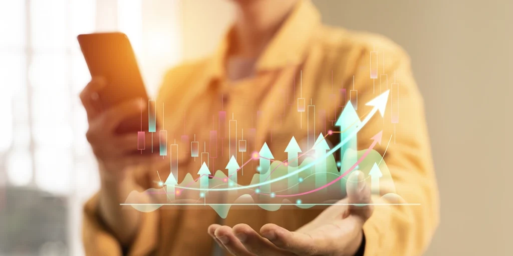 tendências do mercado imobiliário tendências do mercado imobiliário: close na mão de uma mulher. Em cima da mão, vemos um gráfico ascendente com setas indicando para cima. Com a outra mão, a mulher segura um celular.