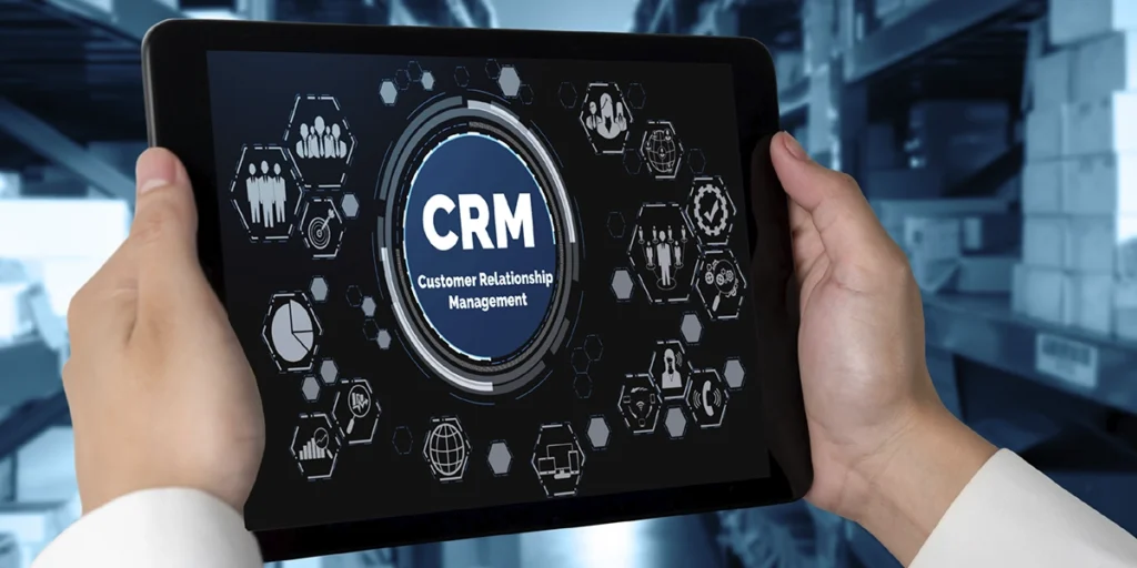 CRM para loteadoras CRM para loteadoras: A imagem mostra uma pessoa segurando uma tablet com a palavra CRM na tela. A pessoa está vestida de branco e preto, e está de pé em um armazém ou instalação de armazéms com estofados e pallets no fundo. A imagem está em preto e branco, com a palavra CRM na tela. A pessoa está segurando a tablet com ambas as mãos, e olha para a tela com uma expressão séria. A imagem é adequada para uso em um contexto empresarial ou tecnológico, e pode ser utilizada para promover um produto ou serviço relacionado à CRM.