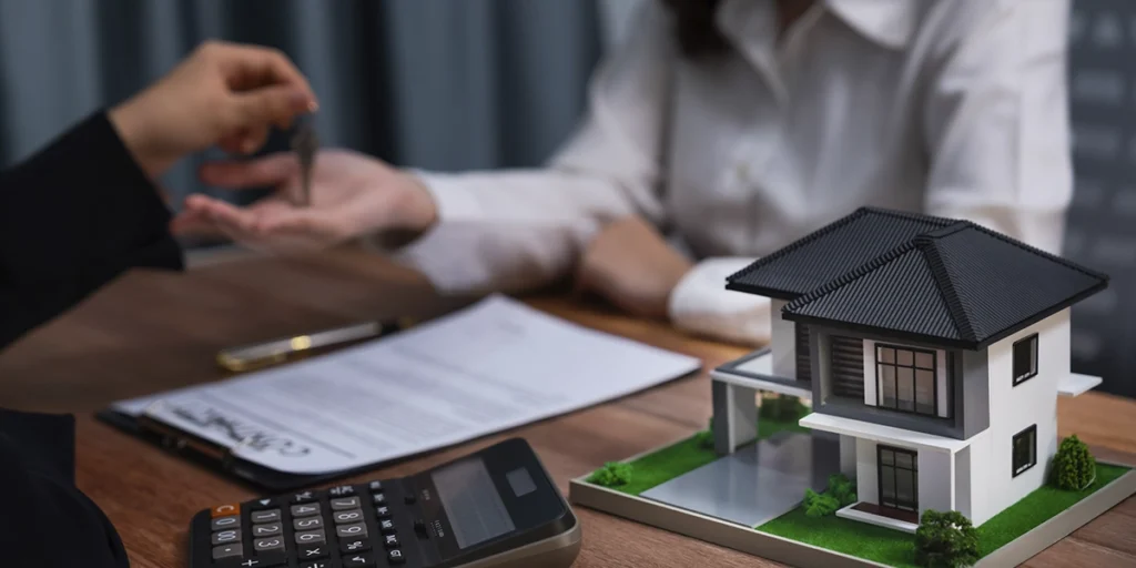 CRM para novos empreendimentos imobiliários CRM para novos empreendimentos imobiliários: close nas mãos de duas pessoas num escritório. Elas estão de lados opostos de uma mesa. Em destaque na imagem, uma casa em miniatura.