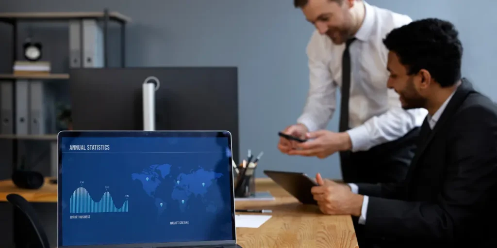 CRM e ERP integrados CRM e ERP integrados: imagem de uma mesa com um notebook em destaque e, ao fundo, dois homens de terno sorriem.